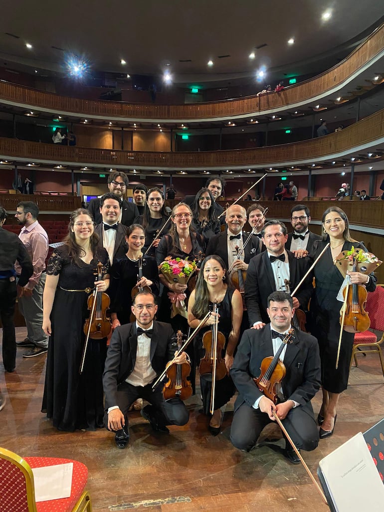 Músicos en traje formal posan en un escenario, una mujer sostiene un ramo de flores en el centro, todos lucen felices.