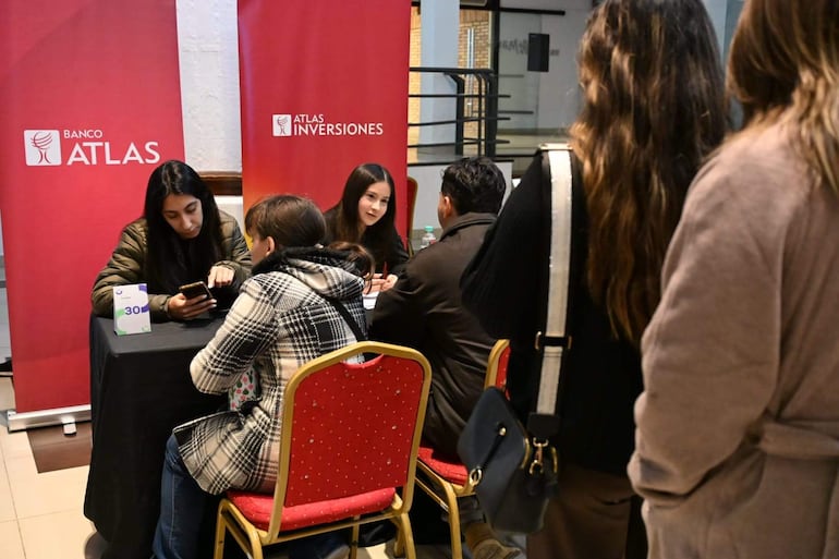 Dos mujeres del Banco Atlas, una usando teléfono, conversan con un hombre en un salón con logos de la institución.