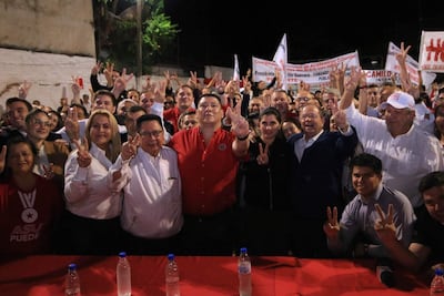 Grupo de personas levantando dos dedos en señal de victoria, vestimenta blanca y roja, sonrisas y ambiente festivo al aire libre.
