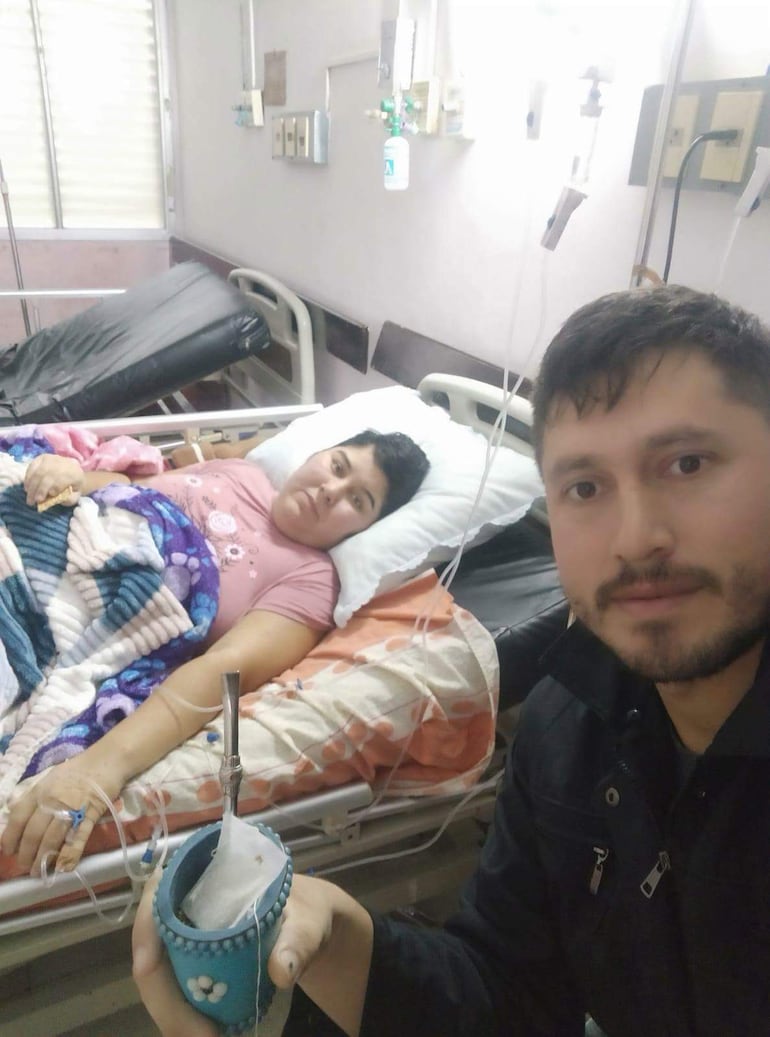 María Ojeda en la cama de un hospital, con manta sobre las piernas, mientras un hombre le toma una selfie con un vaso azul.