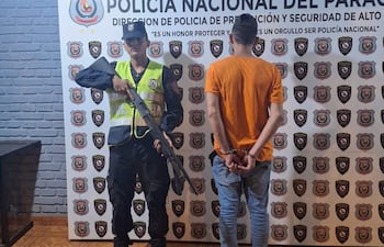 Oficial de policía con arma larga observa a un hombre esposado, ambos en un ambiente interior decorado con insignias policiales.