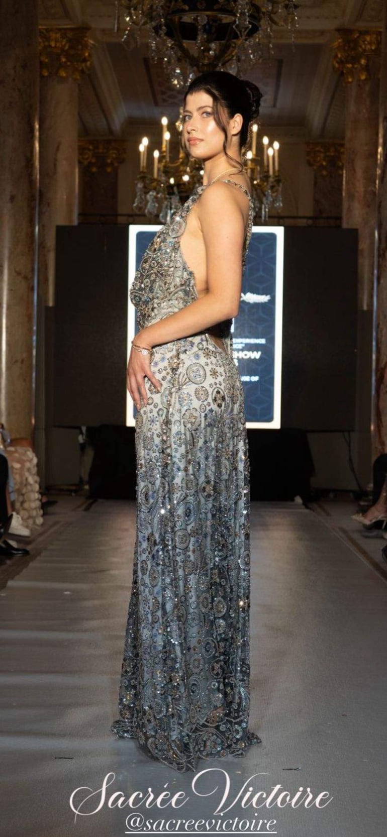 Modelo de perfil en vestido plateado brillante, con expresión serena, rodeada de un ambiente elegante y candelabros.