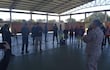 Grupo de personas escucha atentamente a un hombre de pie en uniforme militar y otro con chaqueta negra en un espacio iluminado.