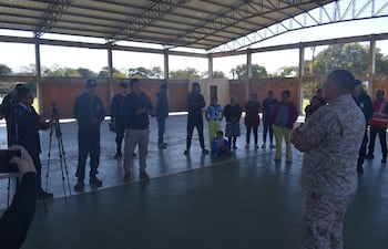 Grupo de personas escucha atentamente a un hombre de pie en uniforme militar y otro con chaqueta negra en un espacio iluminado.