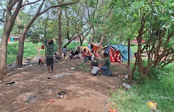 Grupo de personas indígenas en carpas improvisadas, con signos de preocupación y niños en el suelo, rodeados de árboles.