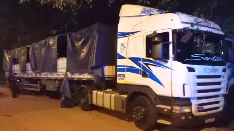 Camión Scania blanco con detalles azules, cubierto con lona negra, estacionado en una calle oscura y desierta.