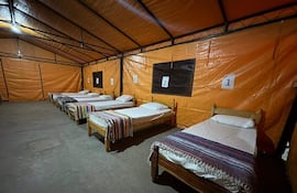 Interior de albergue con seis camas individuales, sábanas blancas y cobertores de colores, ambiente ordenado y sin personas visibles.