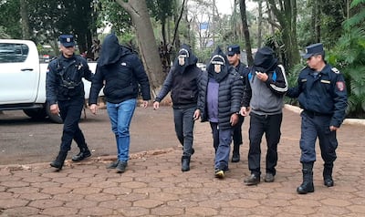 Seis detenidos encapuchados acompañados por un agente policial en un entorno exterior con vegetación al fondo.