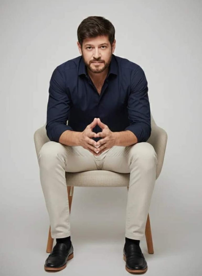Hombre sentado en silla, con camisa oscura y pantalones claros, en un ambiente minimalista y controlado.
