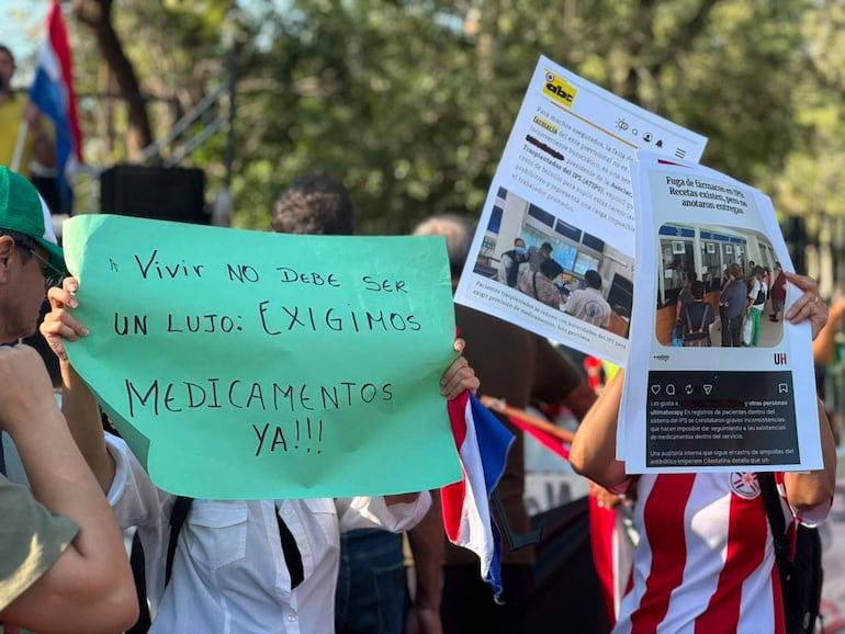 Personas en manifestación sosteniendo carteles. Una persona lleva camiseta a rayas de la Albirroja. Fondo con árboles y bandera paraguaya.
