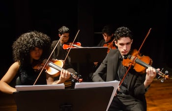 Músicos concentrados en el escenario, con iluminación centrada en ellos y violines en mano, en un ambiente oscuro.