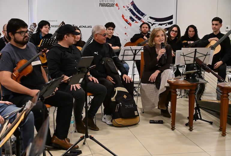 Berta Rojas, con micrófono en mano, rodeada de músicos atentamente escuchando durante la conferencia en un ambiente acogedor.