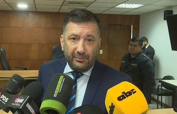 Hombre de mediana edad con barba y traje, rodeado de micrófonos, mientras es entrevistado por la prensa.