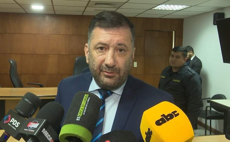 Hombre de mediana edad con barba y traje, rodeado de micrófonos, mientras es entrevistado por la prensa.