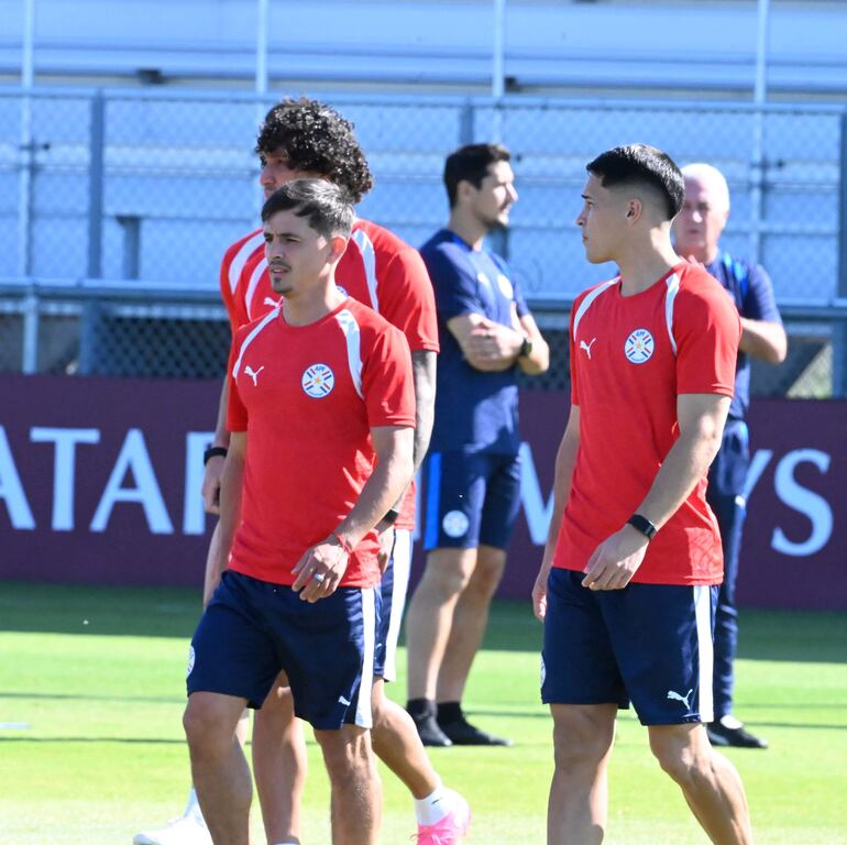 Andrés Cubas con cabello rizado y compañeros en camisetas rojas conversan durante la práctica en un campo de entrenamiento.