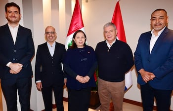 Mujer en blusa oscura interactúa con cuatro hombres, uno alto con traje oscuro y otros con suéteres, rodeados de banderas de Perú y Paraguay.