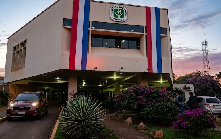 Fachada del Hospital Nacional de Itauguá en tonos rojo, blanco y azul, con vehículos estacionados y cielo anaranjado.