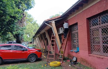 Hombre con camiseta gris y pantalones oscuros sube por una escalera junto a un vehículo rojo, rodeado de vegetación.
