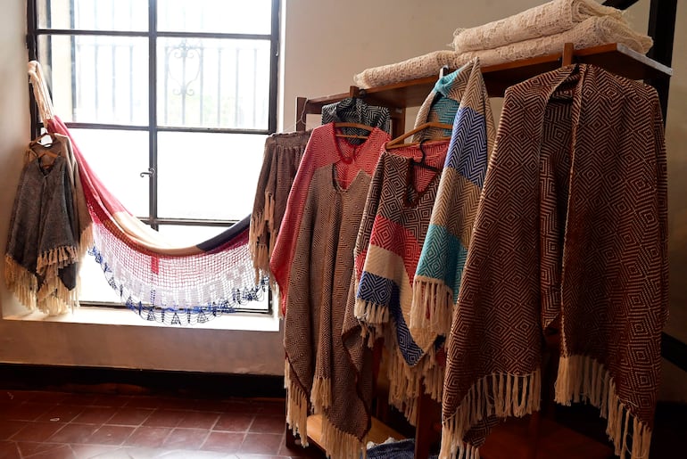 Exhibición de ponchos y mantas en un ambiente cálido y artesanal, con luz natural entrando por una ventana grande.