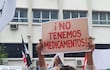 Hombre joven con suéter claro sostiene un cartel en una manifestación. Se observan otros manifestantes y banderas en el fondo.