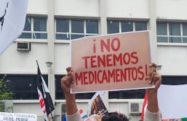 Hombre joven con suéter claro sostiene un cartel en una manifestación. Se observan otros manifestantes y banderas en el fondo.