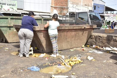 Mujer de espaldas con camiseta azul y pantalones estampados, mientras una niña en camiseta clara observa un contenedor desbordante de basura.