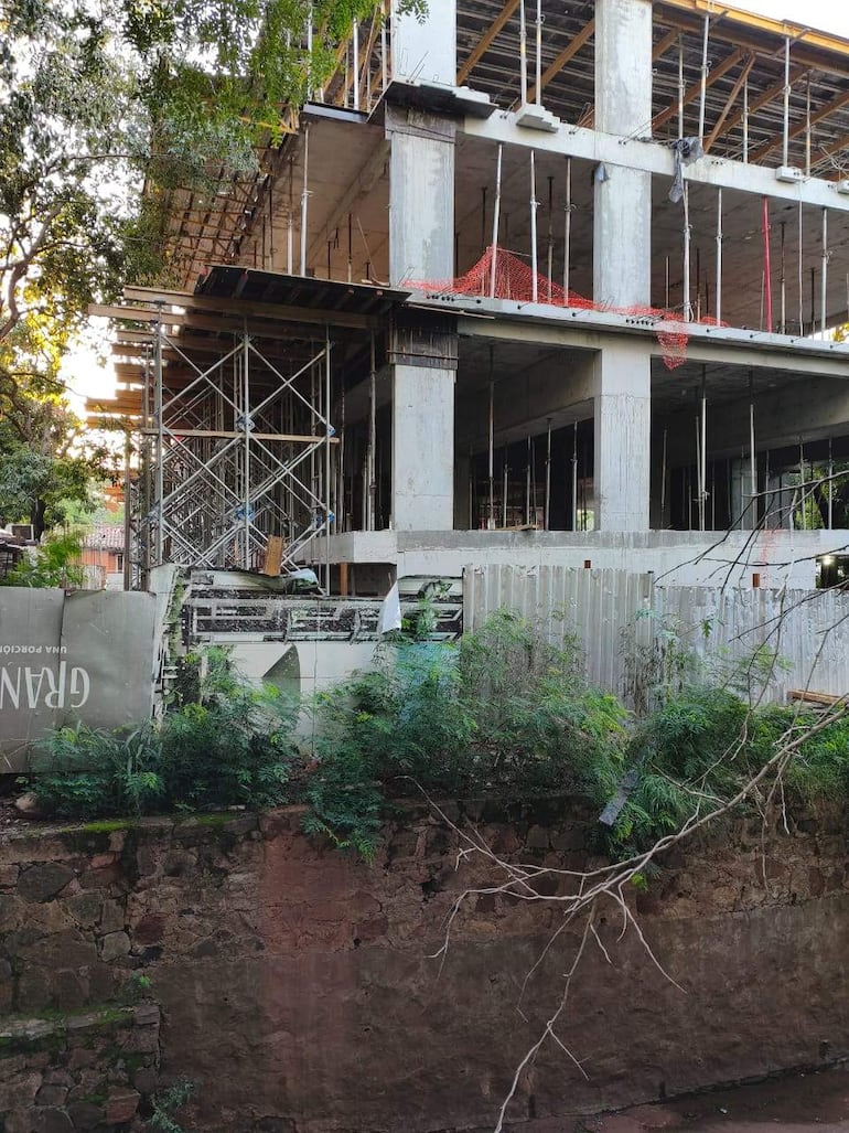 Construcción en proceso con andamiaje y materiales apilados, rodeada de vegetación y un cartel parcialmente visible con la palabra 'GRANO'.