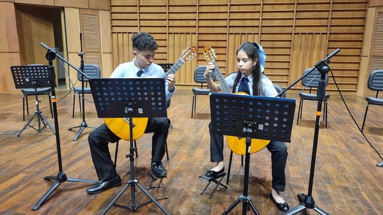 Dos jóvenes con guitarras en tarima, concentrados, vestidos con camisas y corbatas oscuras en un entorno de sala de ensayo.