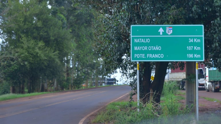 Letrero de dirección verde en la carretera PY07, rodeado de vegetación y camiones en movimiento.