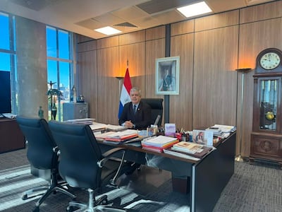 Enrique Riera, hombre canoso en traje oscuro, sentado frente a documentos en oficina luminosa con bandera paraguaya de fondo.