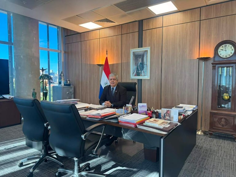 Enrique Riera, hombre canoso en traje oscuro, sentado frente a documentos en oficina luminosa con bandera paraguaya de fondo.