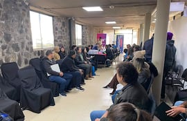 Veinte personas en sala con sillas negras, algunos miran hacia una mesa donde presentan información en un ambiente formal.
