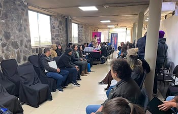Veinte personas en sala con sillas negras, algunos miran hacia una mesa donde presentan información en un ambiente formal.