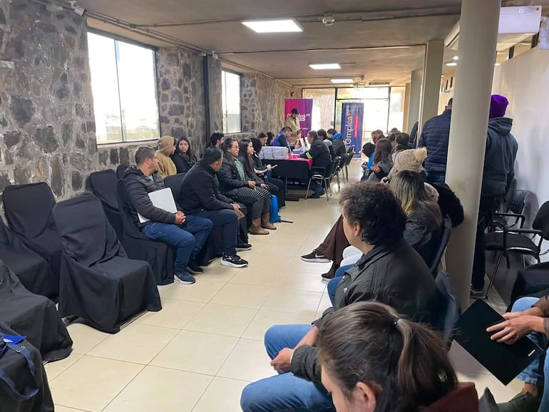 Veinte personas en sala con sillas negras, algunos miran hacia una mesa donde presentan información en un ambiente formal.