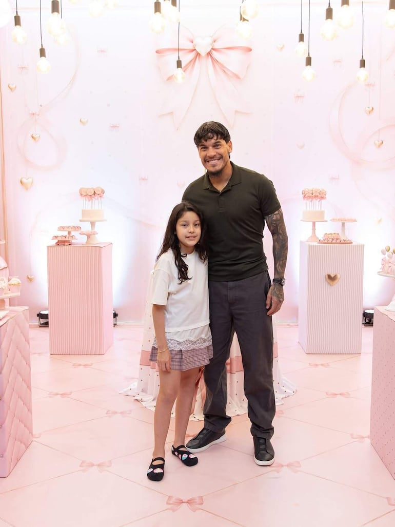 Gustavo Gómez sonríe en camiseta oscura junto a una niña con cabello largo y camiseta blanca, en un entorno festivo con globos rosas.