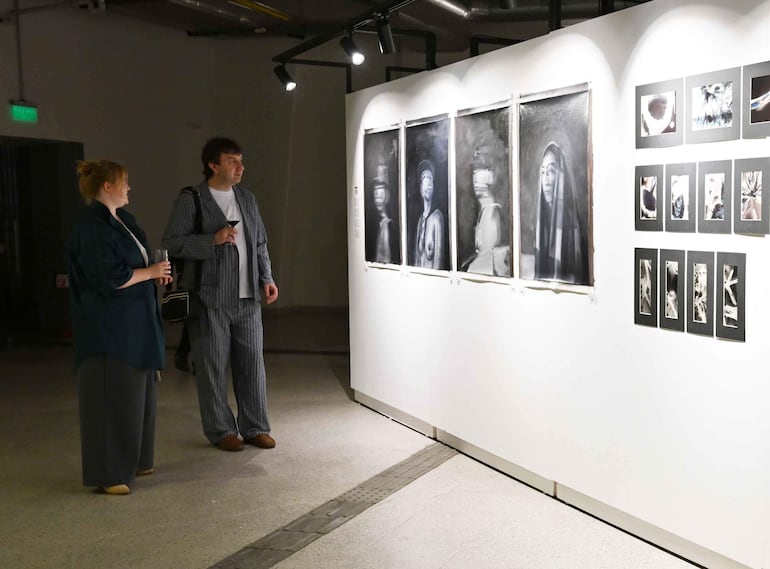 Mujer con blusa oscura y hombre con conjunto a rayas observan obras en galería moderna con iluminación brillante.