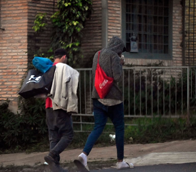 Hombre con mochila roja y abrigo gris camina junto a otro con camiseta negra y abrigo claro por una calle urbana.