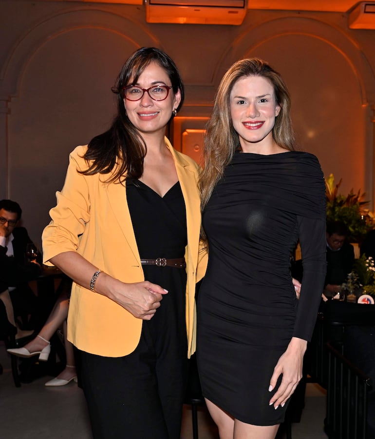 Dos mujeres sonrientes: una con chaqueta amarilla y otra en vestido negro, en un ambiente elegante y festivo.