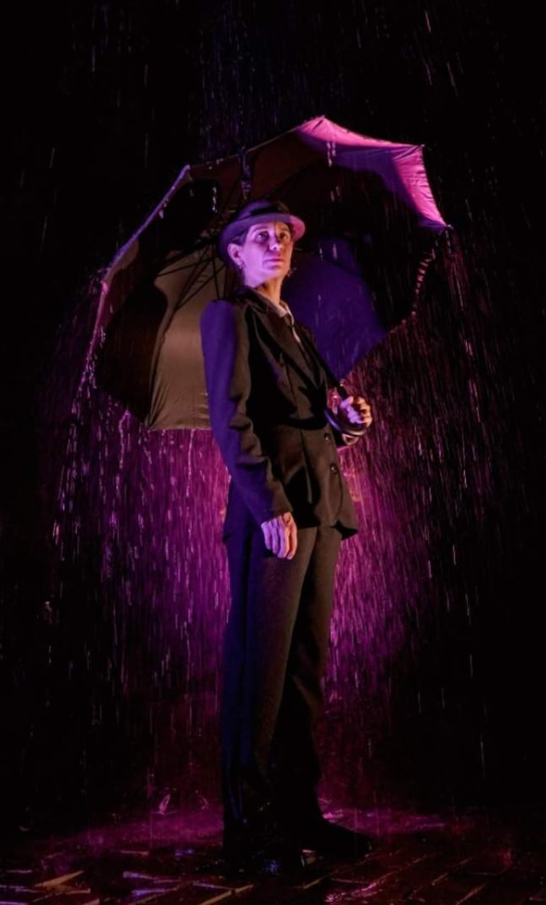 Persona con traje oscuro y paraguas negro bajo lluvia artificial, iluminada con tonos púrpuras, expresión seria y mirada al frente.