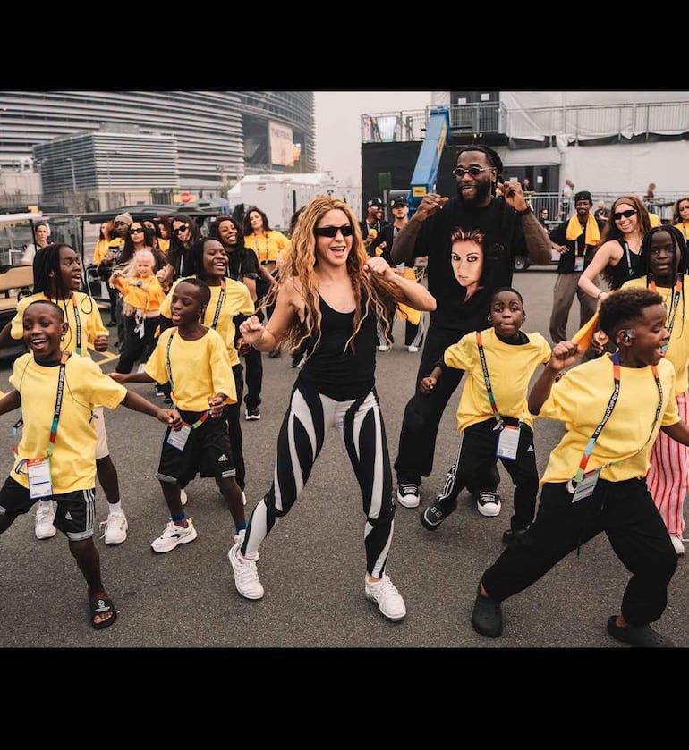 Shakira en camiseta negra bailando con Burna Boy y un grupo de niños con camisetas amarillas, todos lucen felices en un ambiente festivo.