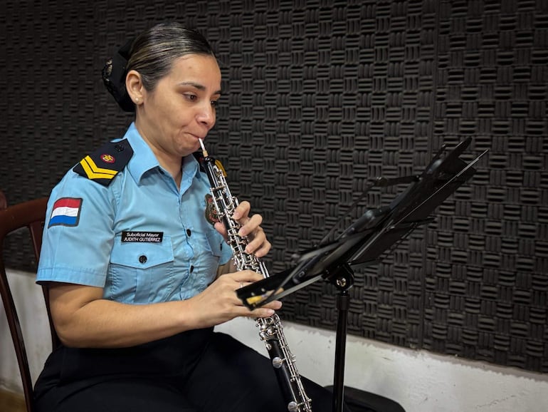 Judith Gutiérrez, vestida con camisa azul y insignias, toca un oboe con expresión concentrada en sala de ensayo.