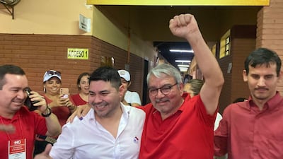 Bernardo Villalba Ayala sonríe y abraza a otros, mientras una mujer con sombrero blanco los observa, todos en un espacio iluminado.
