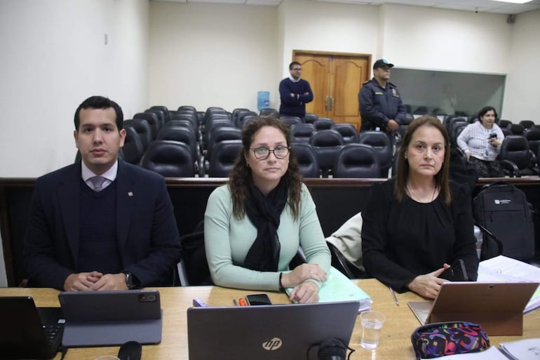 Tres fiscales sentados en mesa. Benítez con camisa y corbata, Guerrero con gafas y cabello rizado, Sánchez con teléfono, en ambiente formal.