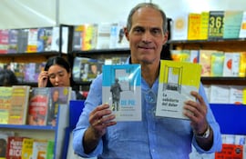Jorge Mendelzon con camisa clara, sonriente, sostiene dos libros frente a estantes llenos de literatura en un ambiente de feria del libro.