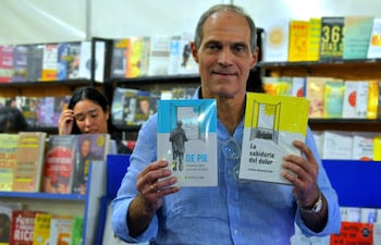 Jorge Mendelzon con camisa clara, sonriente, sostiene dos libros frente a estantes llenos de literatura en un ambiente de feria del libro.
