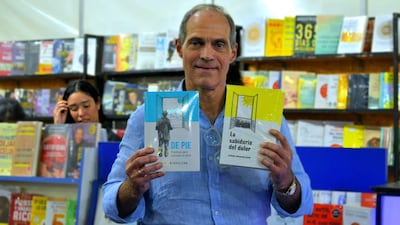 Jorge Mendelzon con camisa clara, sonriente, sostiene dos libros frente a estantes llenos de literatura en un ambiente de feria del libro.