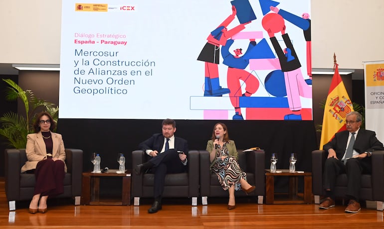 Stella Guillén habla al micrófono, vestida de forma colorida, mientras Fernando Masi y otros la escuchan atentamente en un auditorio moderno.