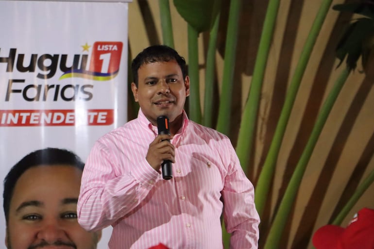 Hugui Farias, con camisa de rayas rosas, habla al público sosteniendo un micrófono, rodeado de plantas decorativas.
