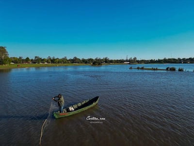 Hombre de pie en canoa verde, usando red de pesca, rodeado de paisaje natural y agua en un día soleado.