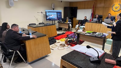 Mujer de pie, con guantes y papeles, presenta pruebas en un tribunal con decoración formal y varias personas atentas en el fondo.